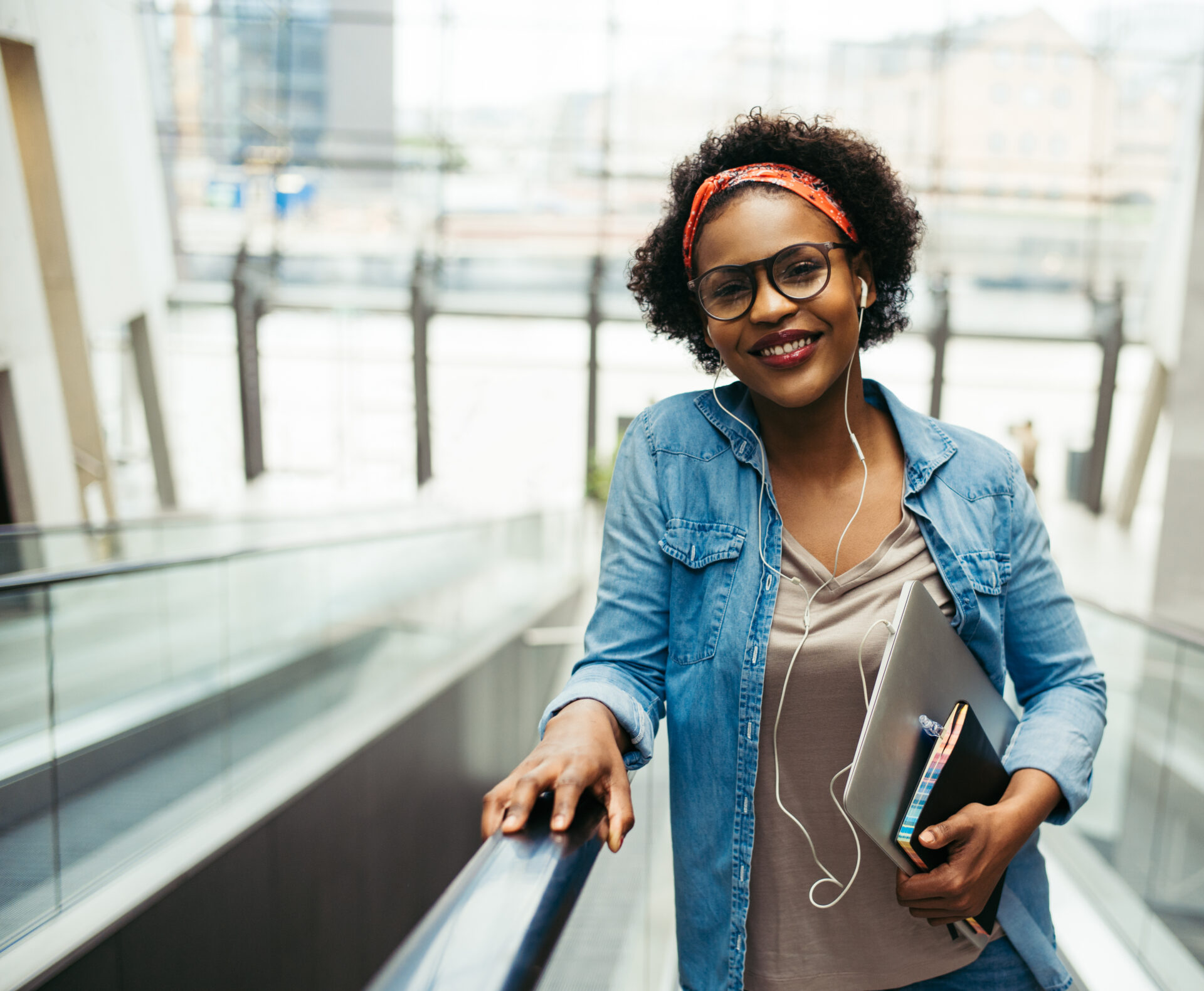 Young African female entrepreneur smiling confidently while riding up an escalator in the lobby of a modern office building carrying a laptop and wearing earphones
